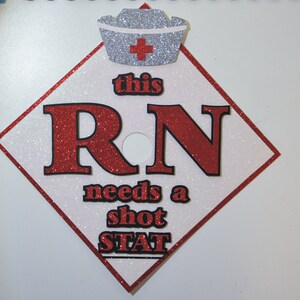 May include: A white diamond-shaped graduation cap with a red border. The cap has a silver glitter nurse's hat with a red cross on top. The text "this RN needs a shot STAT" is written in black and red glitter on the cap.