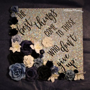 May include: A silver glitter graduation cap decorated with blue, white, and black flowers. The cap has the quote "The best things come to those who don't give up." written in black.