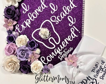 Puede incluir: Un gorro de graduación morado decorado con flores blancas y moradas y un gráfico de diente blanco. El gorro tiene el texto "I explored I scaled I conquered! Deslina" en letras blancas. Un lazo blanco con una flor rosa está unido al gorro con el texto "RDH Nyv 2022 College of Dentistry" en letras negras.