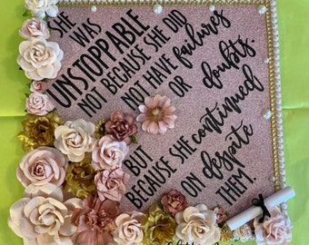 May include: A pink glitter graduation cap decorated with flowers and a quote that reads "She was unstoppable not because she did not have failures or doubts. But because she continued on despite them."