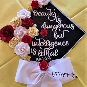 May include: A black graduation cap decorated with white script that reads "Beauty is dangerous but intelligence is lethal." The cap is adorned with red, white, and yellow roses and a white bow. The year "MBA-2021" is also visible.