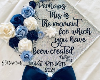 May include: A silver graduation cap decorated with blue and white flowers. The cap has a black glitter background and the text "Perhaps this is the moment for which you have been created. Esther 4:14" is written on it. The cap also has a blue bow and the text "Kat RN BSN 2021" is written on it.