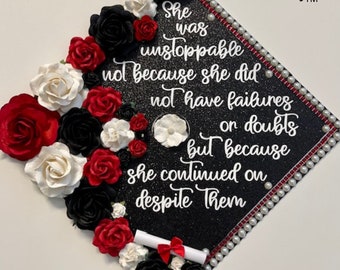 May include: Black graduation cap decorated with red, white, and black paper flowers. The cap has a black glitter background and white pearl trim. The text "She was unstoppable not because she did not have failures or doubts but because she continued on despite them" is written in white on the cap.