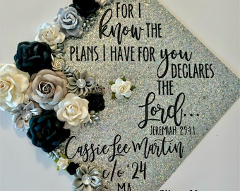 May include: A silver glitter graduation cap decorated with black, white, and gray flowers. The cap has the text "FOR I know THE PLANS I HAVE FOR you DECLARES THE Lord... JEREMIAH 29:11 Cassie Lee Martin 0/0 24 MA GlitterMomz" in black lettering.