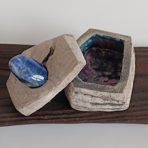 Peut inclure: Une boîte rectangulaire ouverte faite à la main avec un couvercle, fabriquée à partir d'un matériau gris clair. Une pierre bleue repose sur le couvercle. L'intérieur de la boîte est un mélange de bleu et de violet. La boîte repose sur une surface en bois brun foncé.