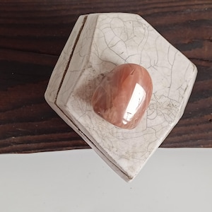 May include: A polished, heart-shaped, orange-brown gemstone sits on a white, diamond-shaped ceramic dish with a crackle glaze. The dish has a brown border and rests on a dark wood surface. The gemstone reflects light.