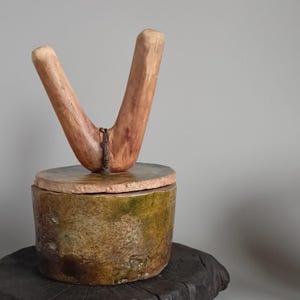Peut inclure: Un récipient cylindrique en céramique avec une glaçure tachetée verte et brune, surmonté d'un couvercle plat et circulaire. Une pièce de bois clair en forme de V est fixée au centre du couvercle par un lien en fil de fer torsadé.