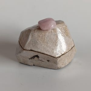 Può includere: Una piccola scatola in ceramica modellata a mano con una forma sfaccettata simile a una roccia e una finitura smaltata color crema con effetto craquelé. Una pietra di quarzo rosa pallido, liscia e lucida, è fissata sul coperchio.