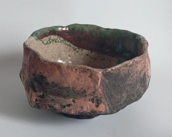 Könnte beinhalten: Eine handgefertigte Keramikschale mit einer rauen, strukturierten Außenseite. Die Schale weist eine Mischung aus Farben auf, darunter Brauntöne, Grün und Schwarz. Der Rand hat eine wellenförmige, organische Form, und das Innere ist hellbeige.