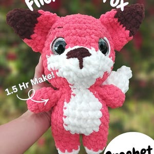 May include: A pink and white crocheted fox toy with large, glittery eyes and brown details. The text "Flicka The Fox" is above the fox, and "1.5 Hr Make!" is on the left. The words "Crochet PDF" are in the bottom right.