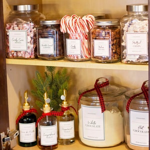 Op de afbeelding: Een glazen kastplank met verschillende glazen potten gevuld met snoep, noten en bakproducten. De potten hebben etiketten met handgeschreven tekst, waaronder "Candy Cove Kisses", "Chocolate Coconut Almonds", "Candy Canes", "Pecan", "Pretzel Crisps", "White Chocolate" en "Hot Chocolate". Er zijn ook vier glazen flessen met tuiten, gelabeld met "Peppermint Syrup", "Gingerbread Syrup" en "Cream of Mint Syrup".