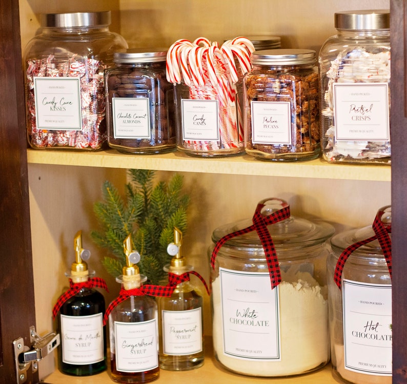 Op de afbeelding: Een glazen kastplank met verschillende glazen potten gevuld met snoep, noten en bakproducten. De potten hebben etiketten met handgeschreven tekst, waaronder "Candy Cove Kisses", "Chocolate Coconut Almonds", "Candy Canes", "Pecan", "Pretzel Crisps", "White Chocolate" en "Hot Chocolate". Er zijn ook vier glazen flessen met tuiten, gelabeld met "Peppermint Syrup", "Gingerbread Syrup" en "Cream of Mint Syrup".