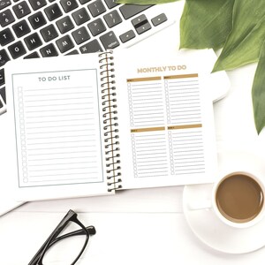 May include: A white notebook with a black spiral binding. The notebook is open to two pages, one labelled "TO DO LIST" and the other labelled "MONTHLY TO DO". The notebook is on a white surface with a laptop computer, a cup of coffee, and a green plant.