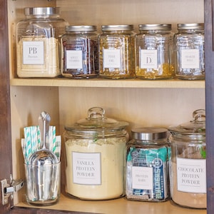 Puede incluir: Un estante de cocina con varios frascos de vidrio que contienen diferentes tipos de proteína en polvo, barras de proteína y suplementos. Los frascos están etiquetados con el tipo de proteína en polvo, como "Proteína en polvo de vainilla", "Proteína en polvo de chocolate" y "Barras de proteína".