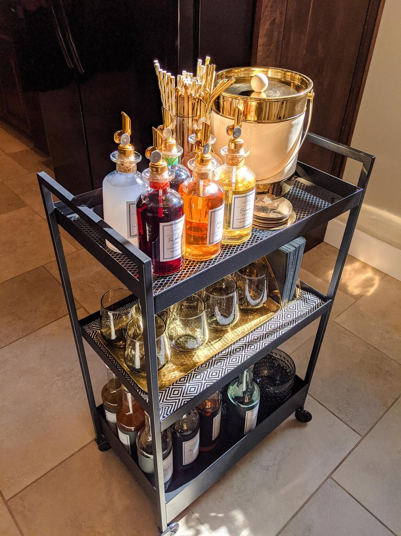 Op de afbeelding: Een zwarte metalen barwagen met twee planken. De bovenste plank heeft een gouden ijs emmer, een bundel gouden rietjes en zes glazen flessen met gouden doppen. De onderste plank heeft een gouden dienblad met zes heldere glazen en zes flessen met gouden doppen.