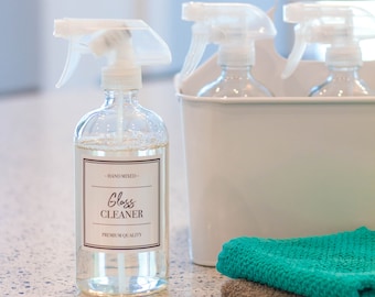 May include: A clear glass bottle with a white label that says "Hand Mixed - Glass Cleaner - Premium Quality". The bottle has a white spray nozzle and is sitting on a white countertop. There are two other bottles with white spray nozzles in a white bucket behind the bottle.