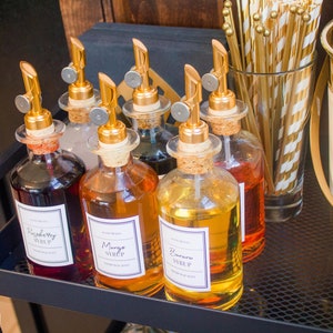 Op de afbeelding: Vijf glazen flessen met gouden doppen en etiketten op een zwart metalen rek. De etiketten lezen "Frambozensiroop", "Mangosiroop", "Bananen siroop", "Vanillesiroop" en "Chocoladesiroop".
