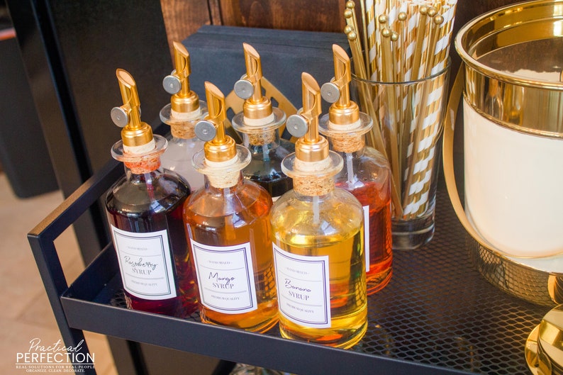 Op de afbeelding: Vijf glazen flessen met gouden doppen en etiketten op een zwart metalen rek. De etiketten lezen "Frambozensiroop", "Mangosiroop", "Bananen siroop", "Vanillesiroop" en "Chocoladesiroop".