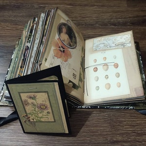 May include: Open journal with vintage-style pages, including botanical illustrations and a portrait. A black frame with a floral print is in the foreground. The journal is bound with a black ribbon and has metal corner protectors.