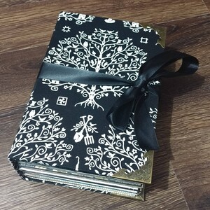 May include: A black and white fabric-covered journal with a tree of life design and brass corner protectors. The journal is tied with a black satin ribbon. The cover features a repeating pattern of trees, birds, and garden tools.