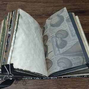 May include: An open journal with aged paper pages, held together with binder clips and a black ribbon. The pages include printed vintage newspaper clippings and decorative patterned paper. The journal's cover has metal corner protectors.