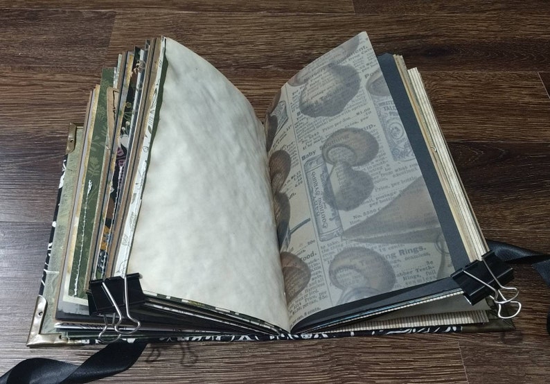 May include: An open journal with aged paper pages, held together with binder clips and a black ribbon. The pages include printed vintage newspaper clippings and decorative patterned paper. The journal's cover has metal corner protectors.