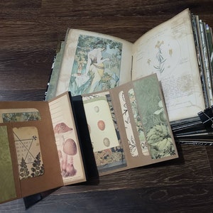 May include: Open journal with botanical illustrations and decorative inserts. The journal features a variety of pages, including lined paper, botanical prints, and pockets. A black binder clip holds the pages together. The journal is open on a dark wood surface.