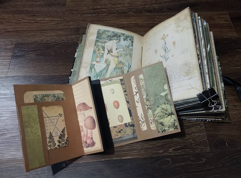 May include: Open journal with botanical illustrations and decorative inserts. The journal features a variety of pages, including lined paper, botanical prints, and pockets. A black binder clip holds the pages together. The journal is open on a dark wood surface.