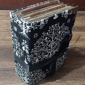 May include: A black and white fabric-covered journal with a tree of life design. The journal is bound with a black ribbon and has metal corner protectors. The pages are visible and appear to be aged.