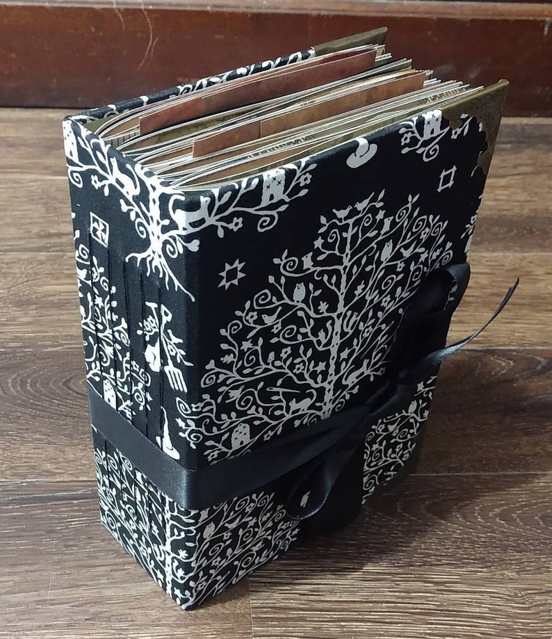 May include: A black and white fabric-covered journal with a tree of life design. The journal is bound with a black ribbon and has metal corner protectors. The pages are visible and appear to be aged.