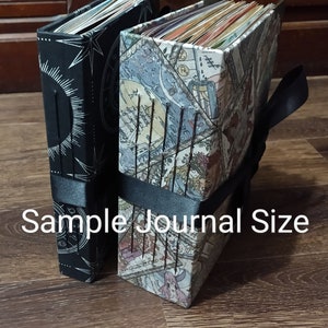 May include: Two journals with decorative covers. One is black with a white sunburst design, the other has a colorful map-like pattern. Both are bound with black ribbon. The text "Sample Journal Size" is visible.