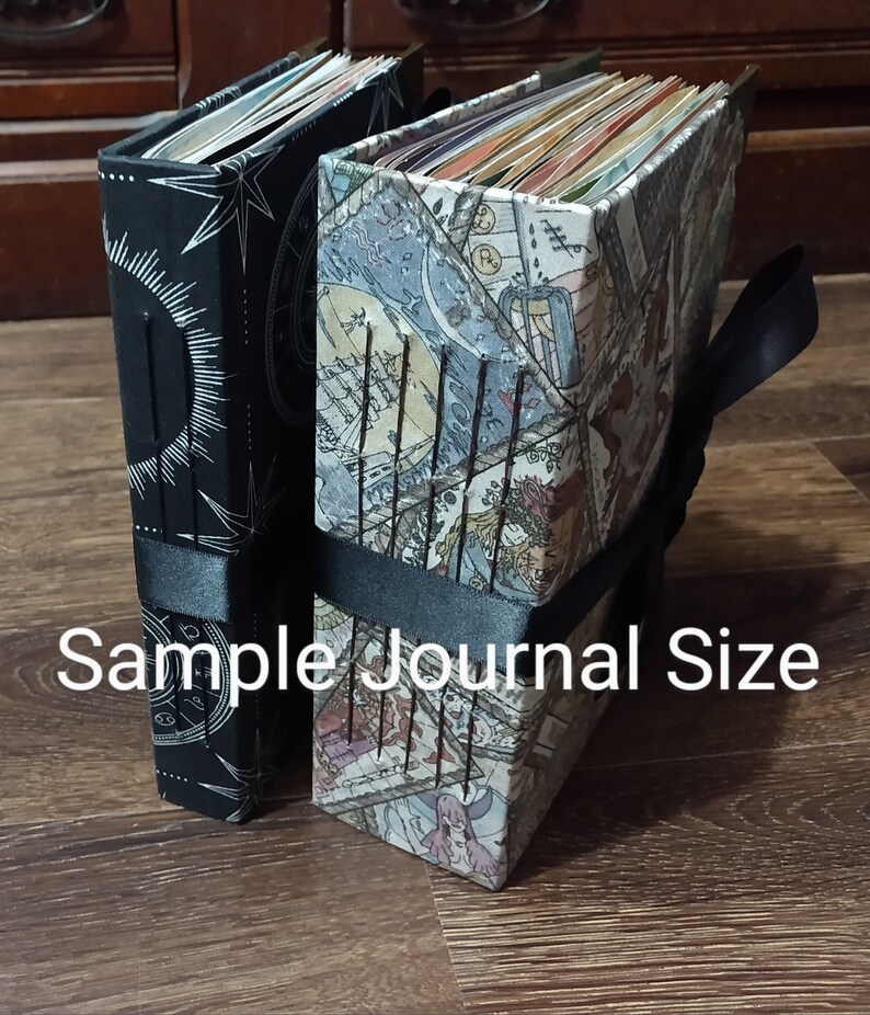 May include: Two journals with decorative covers. One is black with a white sunburst design, the other has a colorful map-like pattern. Both are bound with black ribbon. The text "Sample Journal Size" is visible.