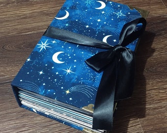 Könnte beinhalten: Ein Hardcover-Journal mit einem Himmelsdesign. Der Einband hat einen dunkelblauen Hintergrund mit weißen Monden und Sternen. Ein schwarzes Satinband ist um das Journal gebunden. Die Ecken sind mit goldfarbenem Metall geschützt.