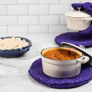 May include: A white enameled cast iron pot with a lid, filled with stew, sits on a purple patterned trivet. A matching pot is held by a person wearing purple oven mitts. A blue bowl of rice is in the background.