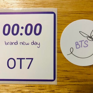 May include: Two stickers, one with a purple border and the text "00:00 brand new day OT7" and the other with a butterfly design and the text "BTS" in purple.