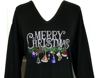 Puede incluir: Camiseta negra de manga larga con cuello en V. La parte delantera presenta el texto "MERRY CHRISTMAS" en pedrería blanca, junto con adornos navideños. Los adornos incluyen un regalo, un bastón de caramelo, un calcetín y un gorro de Papá Noel.