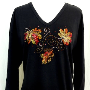 Könnte beinhalten: Schwarzes Langarmshirt mit V-Ausschnitt. Das Shirt hat ein Design aus Herbstblättern in Rot-, Orange- und Gelbtönen, verziert mit glitzernden Strasssteinen. Die Blätter sind mit einem wirbelnden Rankenmuster angeordnet.