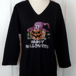 May include: Black long-sleeve shirt with a V-neck. The shirt features a rhinestone design of a jack-o'-lantern wearing a witch hat. The words "HAPPY HALLOWEEN" are spelled out in rhinestones below the pumpkin.