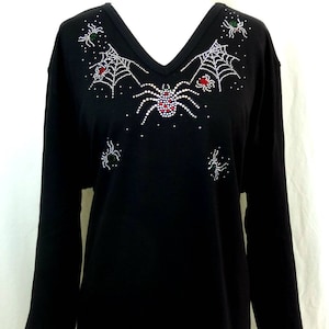 Peut inclure: T-shirt noir &agrave; manches longues et col en V. Le t-shirt est orn&eacute; d'un motif de toile d'araign&eacute;e et d'araign&eacute;es en strass blancs, rouges et verts. Le motif est centr&eacute; sur la poitrine et pr&egrave;s de l'encolure.
