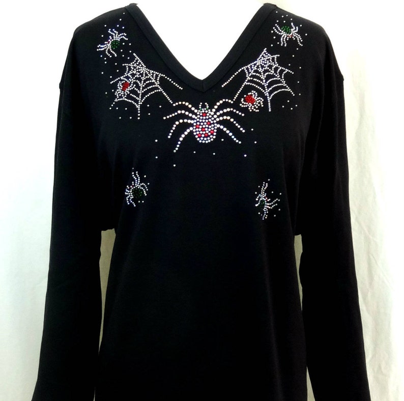 Peut inclure: T-shirt noir &agrave; manches longues et col en V. Le t-shirt est orn&eacute; d'un motif de toile d'araign&eacute;e et d'araign&eacute;es en strass blancs, rouges et verts. Le motif est centr&eacute; sur la poitrine et pr&egrave;s de l'encolure.