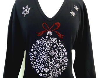 Puede incluir: Camiseta negra de manga larga con un diseño de adorno navideño de strass rojo y blanco. El adorno está decorado con copos de nieve y un lazo rojo.