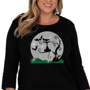 May include: Black long-sleeved shirt with a rhinestone design of a black cat sitting on a tree branch in front of a full moon. The moon and tree are outlined with rhinestones, and there are two bats flying in the air. The shirt has a green grass design at the bottom.