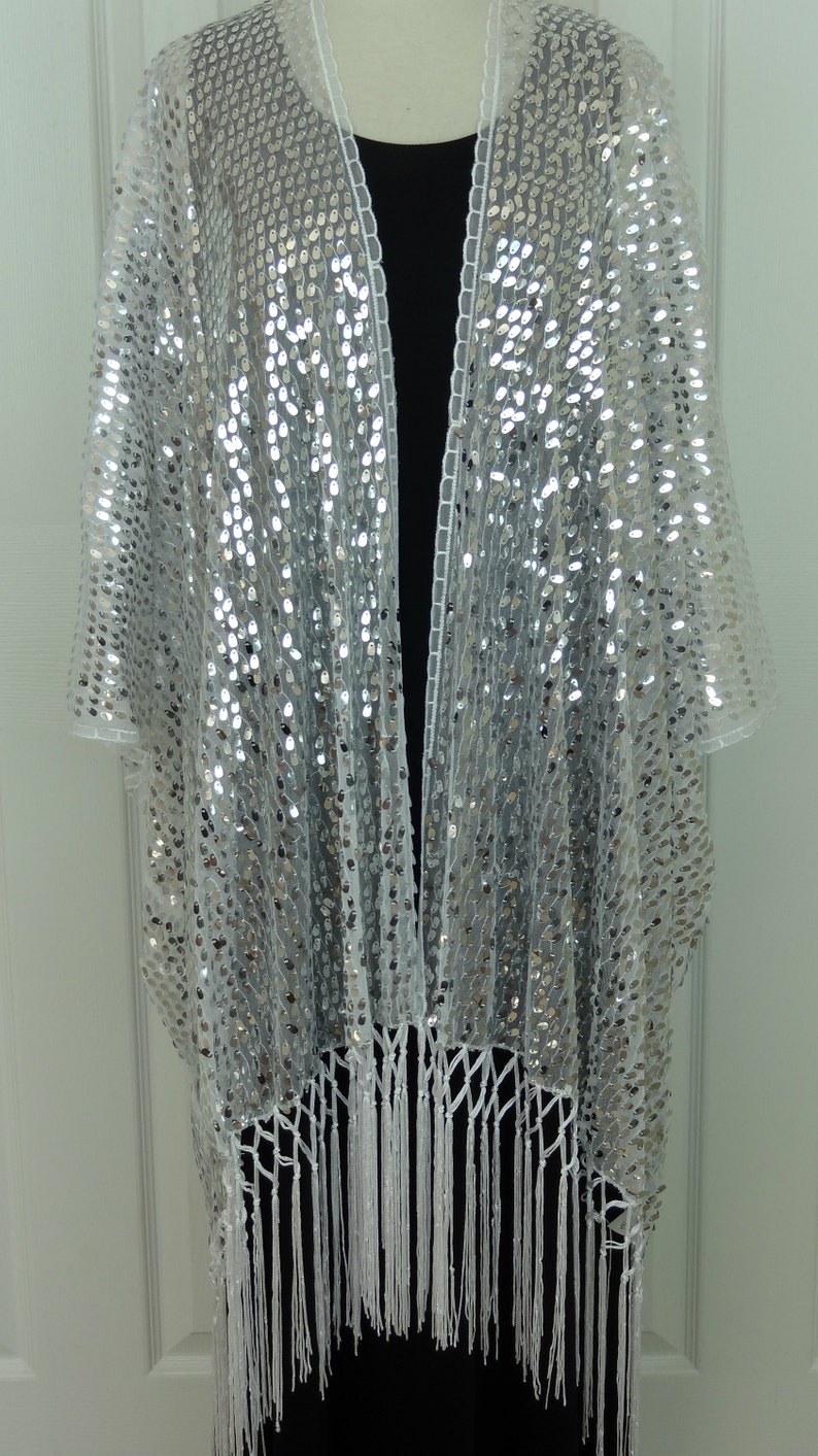 May include: A white sequined shawl with a fringe border. The shawl is open in the front and has a V-neckline.