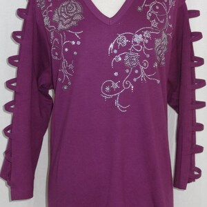 May include: A long-sleeved, purple t-shirt with a v-neck and a rhinestone floral design. The sleeves have cutouts along the sides.