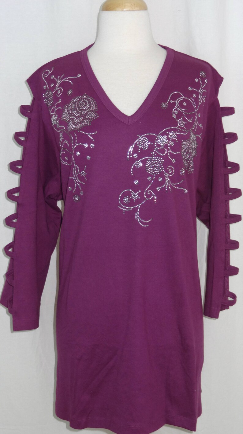 May include: A long-sleeved, purple t-shirt with a v-neck and a rhinestone floral design. The sleeves have cutouts along the sides.