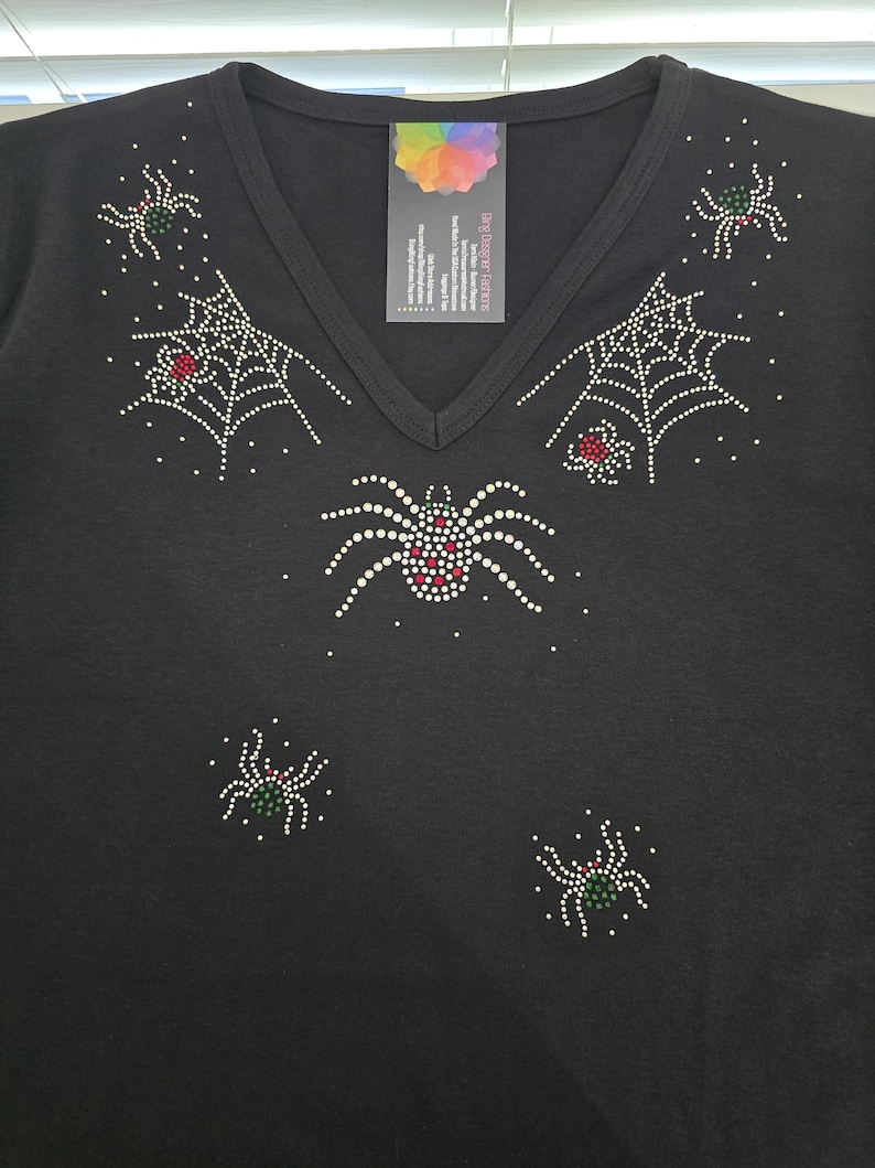 Peut inclure: T-shirt noir &agrave; col en V orn&eacute; de motifs d'araign&eacute;es et de toiles d'araign&eacute;es en strass. Les araign&eacute;es ont des accents rouges et verts. Une &eacute;tiquette avec un logo color&eacute; est attach&eacute;e &agrave; l'encolure. V&ecirc;tement sur le th&egrave;me d'Halloween ou gothique.