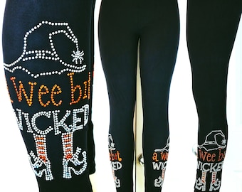 Puede incluir: Leggings negros con diseño de pedrería. El diseño incluye un sombrero de bruja, una araña y las palabras "a Wee bit Wicked" en pedrería naranja y blanca. Un artículo temático de Halloween.