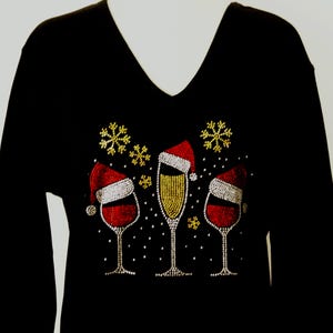 Pode incluir: Camiseta preta de manga comprida com decote em V com um design festivo. O design apresenta três taças de vinho, duas cheias de vinho tinto e uma de champanhe, todas usando chapéus de Papai Noel. Flocos de neve dourados e pontos brancos completam o tema natalino.