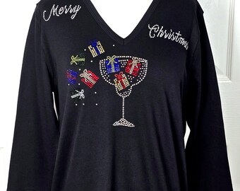 Puede incluir: Camiseta negra de manga larga con cuello en V y las palabras "Merry Christmas" en pedrería. La camiseta presenta un diseño de una copa de vino con regalos que se derraman de ella, también hechos de pedrería.