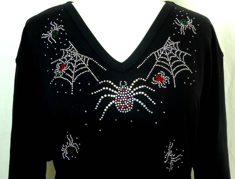 Peut inclure: T-shirt noir &agrave; manches longues et col en V, orn&eacute; de motifs d'araign&eacute;es et de toiles d'araign&eacute;es en strass. Les araign&eacute;es ont des d&eacute;tails rouges, verts et blancs. Le motif est centr&eacute; sur la poitrine et les &eacute;paules, cr&eacute;ant un th&egrave;me d'Halloween.
